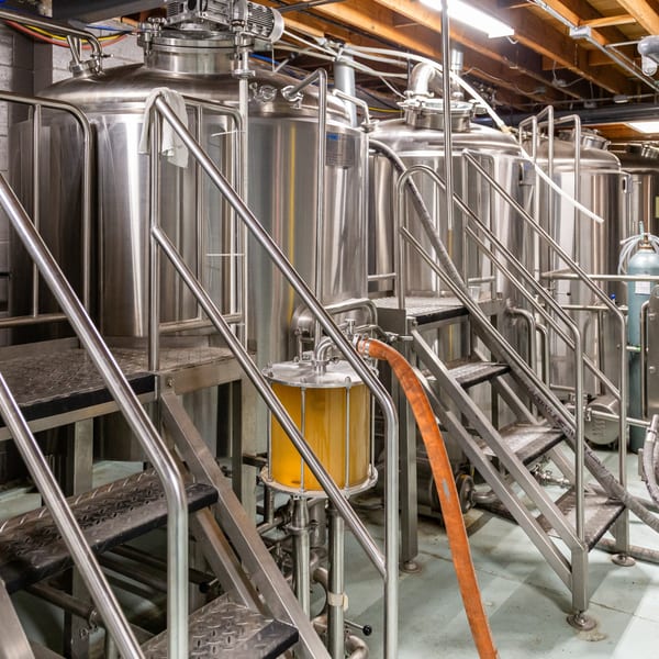 Stainless steel brewing tanks and equipment with stairs, hoses, and a clear vessel showing amber liquid.