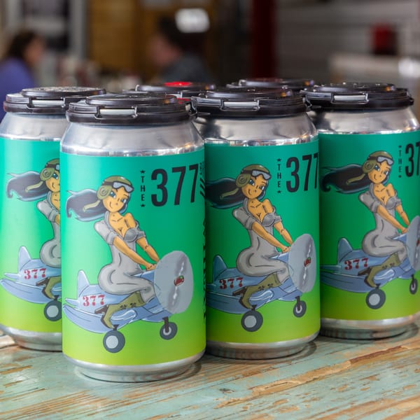 Several 377 Brewery beer cans with a pin-up girl riding an airplane design on a wooden table.