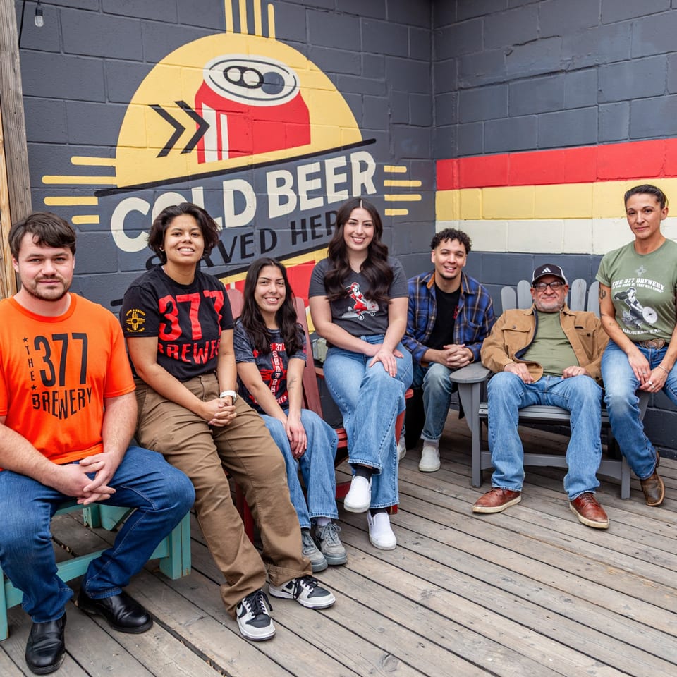 Seven people, some wearing 377 Brewery shirts, sit on benches in front of a colorful brewery mural.