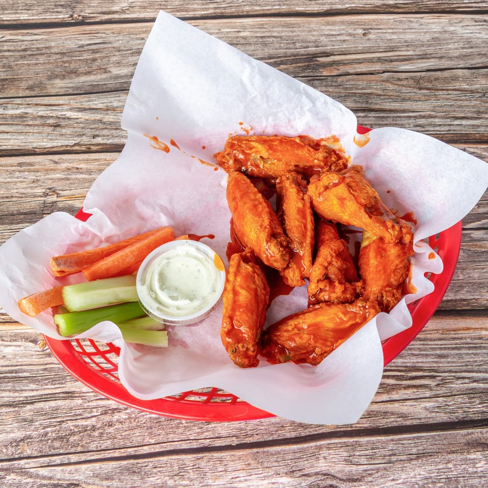 A red basket of buffalo wings with carrots, celery, and ranch dressing on a wooden table.