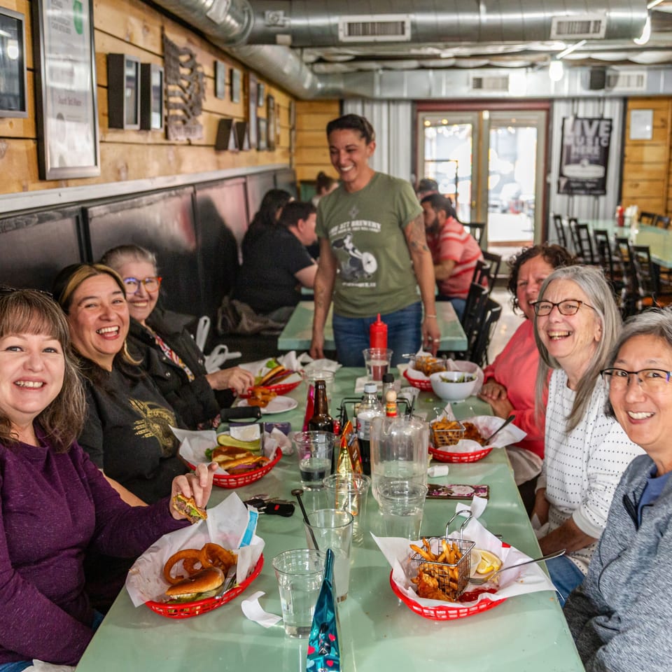 Smiling patrons and a server at The 377 Brewery, enjoying burgers and fries at a long table.