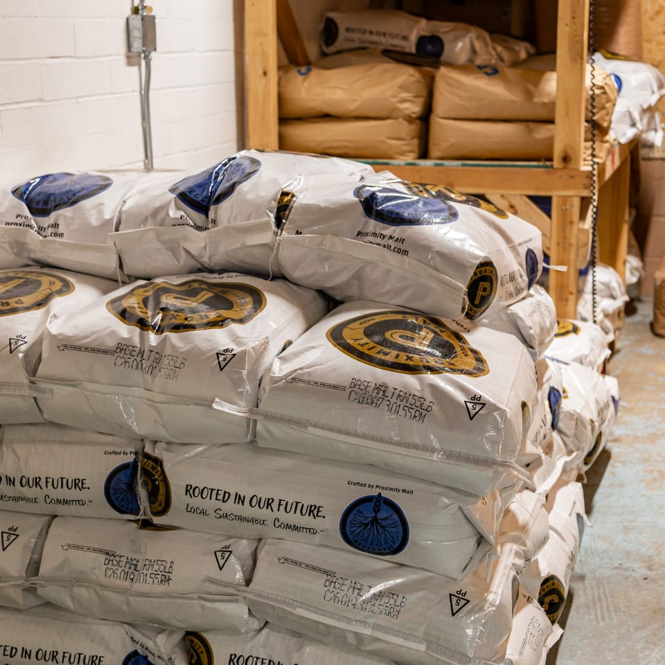 Stacked bags of "Proximity Malt" in a brewery storage area, ready for brewing.