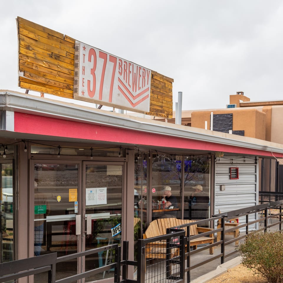 Exterior of The 377 Brewery with a wooden sign, red awning, and glass doors on a cloudy day.