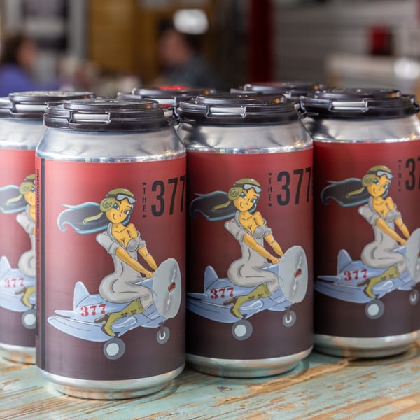 Several 377 Brewery beer cans with pin-up girl and airplane art on a wooden table.
