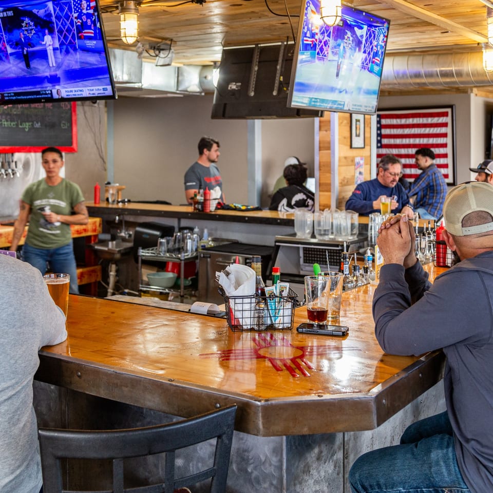 Patrons and a bartender at a wooden bar with TVs showing sports, American flag in background.