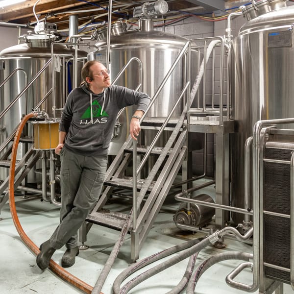 A man in a Haas hoodie leans on a metal staircase in a brewery with large stainless steel tanks.