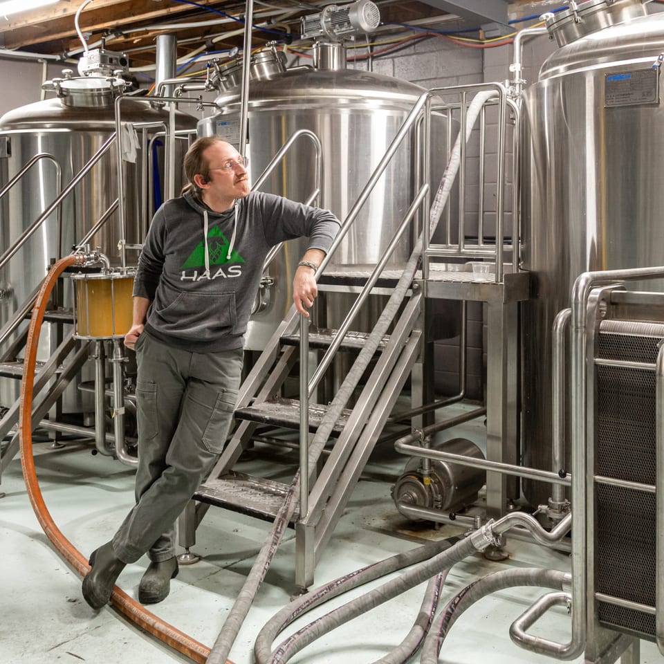 A man in a Haas hoodie leans on a metal staircase in a brewery with large stainless steel tanks.