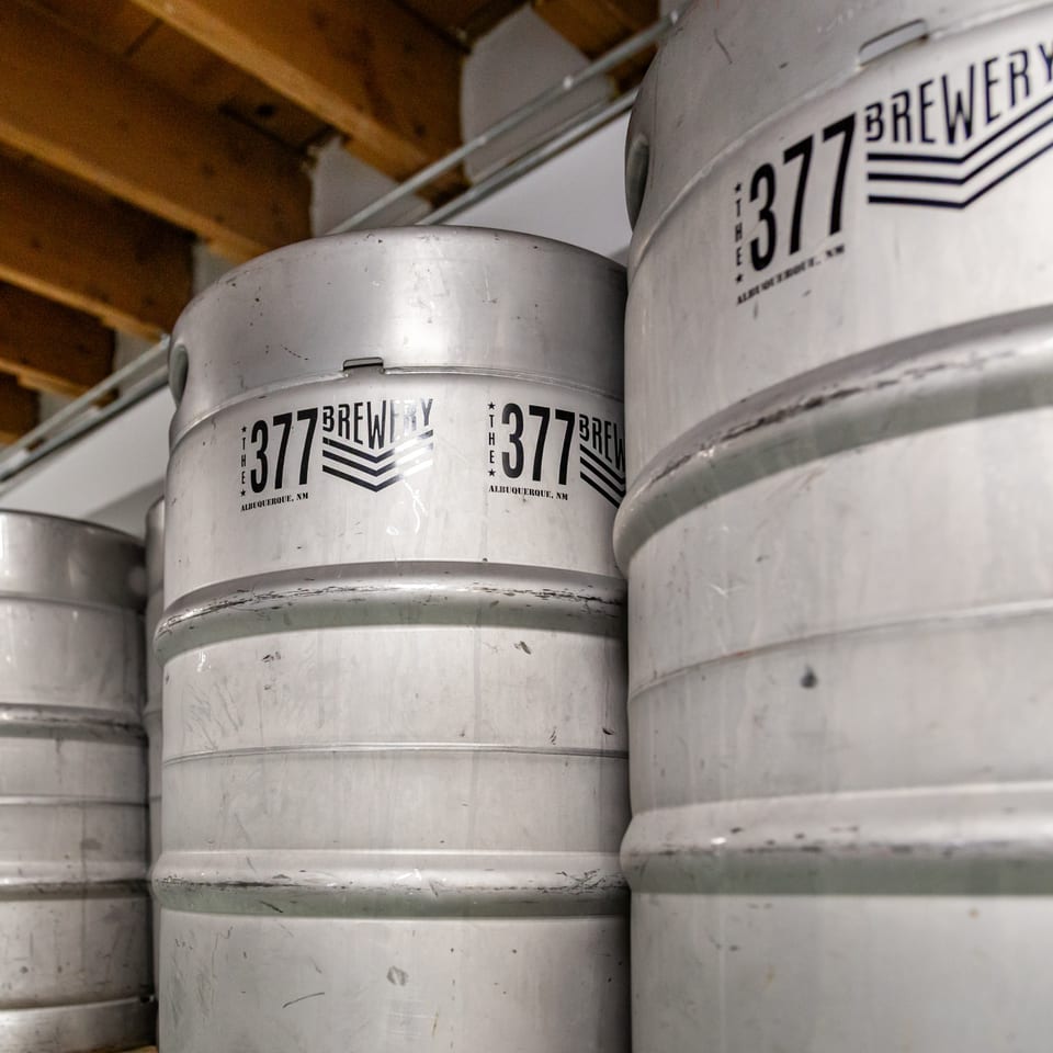 Several silver beer kegs with "The 377 Brewery Albuquerque, NM" logo are stacked in a storage area.