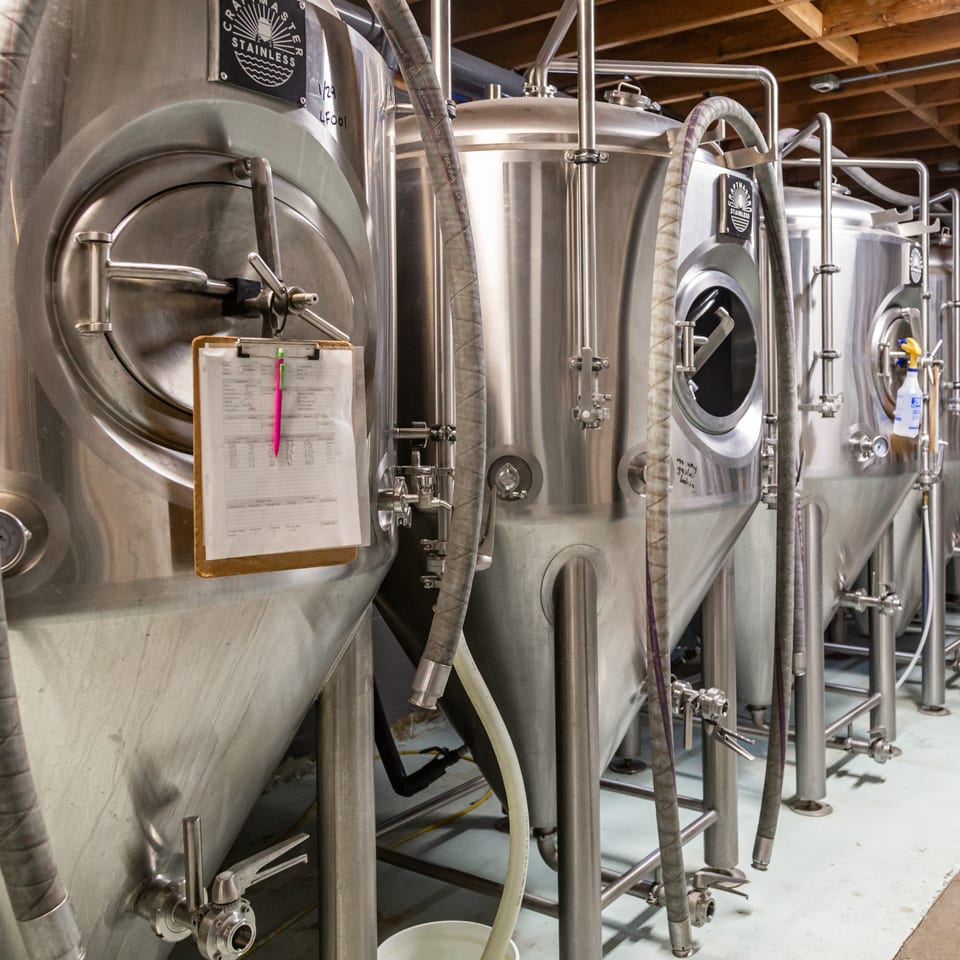 Row of large stainless steel fermentation tanks with hoses and a clipboard at The 377 Brewery.