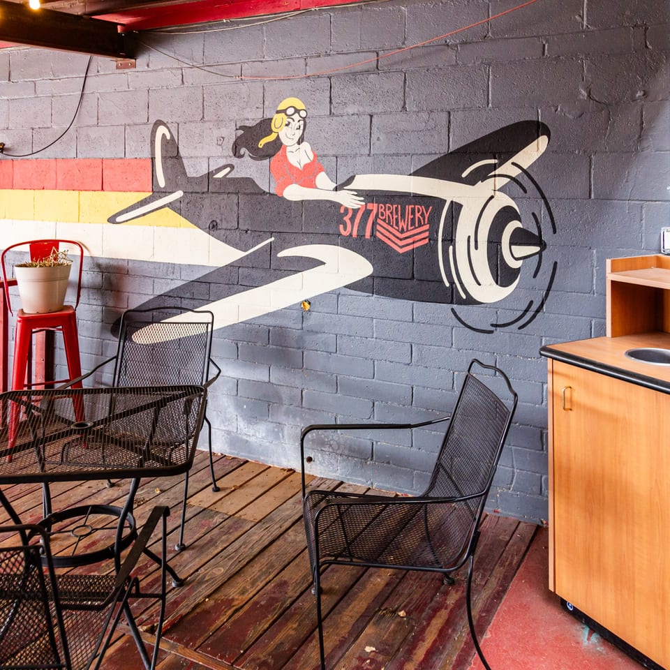 A mural of a female pilot on a plane with "377 Brewery" on a brick wall, with outdoor seating.
